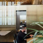 A person works alone at a laptop in an elegant open-plan office with glass walls and ornate architectural ceiling details