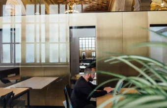 A person works alone at a laptop in an elegant open-plan office with glass walls and ornate architectural ceiling details