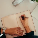 A person's hand writes in an open notebook next to a laptop keyboard on a clean desk, representing writing persistent instructions for an AI coding assistant