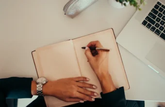 A person's hand writes in an open notebook next to a laptop keyboard on a clean desk, representing writing persistent instructions for an AI coding assistant