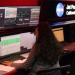 A NASA Deep Space Network operations chief monitors real-time telemetry and status dashboards across multiple screens at the Jet Propulsion Laboratory