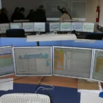A bank of computer monitors displaying system status screens at a control room operations desk, illustrating console sprawl in IT operations