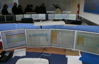 A bank of computer monitors displaying system status screens at a control room operations desk, illustrating console sprawl in IT operations