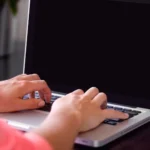 Close-up of two hands typing on a laptop keyboard with a dark blank screen