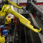 Two yellow FANUC industrial robot arms positioned over a steel beam on a factory floor, preparing to weld