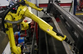 Two yellow FANUC industrial robot arms positioned over a steel beam on a factory floor, preparing to weld