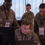 Soldiers in camouflage uniforms gather around a laptop and monitor, reviewing network activity during a cyber defense training exercise