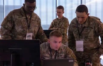 Soldiers in camouflage uniforms gather around a laptop and monitor, reviewing network activity during a cyber defense training exercise