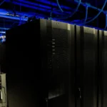 Rows of dark data center server racks in a corridor lit by blue overhead liquid cooling infrastructure