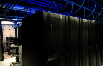 Rows of dark data center server racks in a corridor lit by blue overhead liquid cooling infrastructure