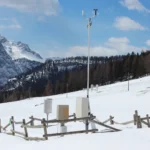 An automated weather station with wind speed and temperature sensors mounted on a pole in a snowy alpine landscape in the Austrian Alps, with mountains in the background