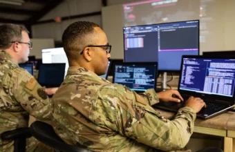 Military cyber operators monitor several computer screens showing code and network data during a cybersecurity exercise