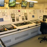 A ship engine control room with rows of monitors, gauges, and control panels, an empty operator chair in the foreground