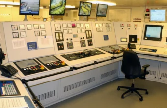A ship engine control room with rows of monitors, gauges, and control panels, an empty operator chair in the foreground