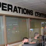 Airmen sit at computer monitors below an OPERATIONS CENTER sign, with wall-mounted status screens showing tracked metrics behind them