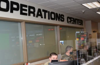 Airmen sit at computer monitors below an OPERATIONS CENTER sign, with wall-mounted status screens showing tracked metrics behind them