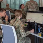 Two soldiers analyze defensive data logs together on a computer terminal, one pointing at a line on screen, representing how the right context changes what an AI agent can diagnose