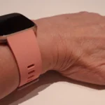 A Fitbit Versa smartwatch with a pink band worn on a wrist, screen off, resting on a light surface