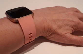 A Fitbit Versa smartwatch with a pink band worn on a wrist, screen off, resting on a light surface