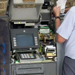 A technician's hand reaches into an open ATM, revealing exposed circuit boards, a screen, and a keypad above the machine's locked cash safe