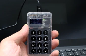A hand holds a Coinkite Coldcard hardware wallet showing a Bitcoin transaction confirmation screen and numeric keypad
