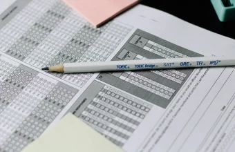 A pencil labeled with standardized test names rests across a multiple-choice answer sheet, evoking the exam analogy behind double-blind AI evaluation