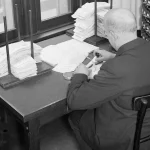 Black and white 1933 photo of an office clerk stamping documents at a desk, surrounded by two large rotating racks holding hundreds of labeled rubber stamps