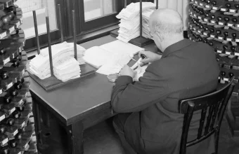 Black and white 1933 photo of an office clerk stamping documents at a desk, surrounded by two large rotating racks holding hundreds of labeled rubber stamps