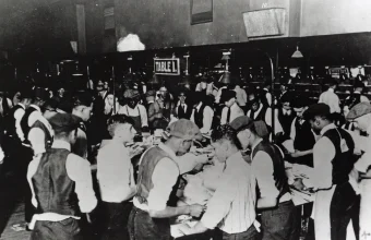 Black and white 1890s photo of postal clerks sorting mail by hand at a crowded sorting table, pigeonhole shelving visible in the background