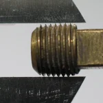 A caliper's jaws holding a brass threaded pipe plug against a plain background, checking whether it conforms to a fixed size