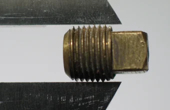 A caliper's jaws holding a brass threaded pipe plug against a plain background, checking whether it conforms to a fixed size