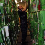 A U.S. Navy sailor in uniform reaches up to file a green medical record folder among long rows of shelved patient records in a hospital records room.