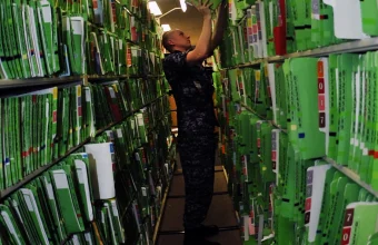 A U.S. Navy sailor in uniform reaches up to file a green medical record folder among long rows of shelved patient records in a hospital records room.