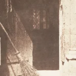 1844 sepia photograph titled The Open Door by William Henry Fox Talbot, showing a rustic open wooden doorway with a broom leaning against it and a lantern on the wall