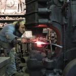 A blacksmith uses an industrial power hammer to forge glowing hot steel, illustrating Docker's push to harden software down to the OS package level