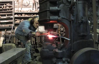 A blacksmith uses an industrial power hammer to forge glowing hot steel, illustrating Docker's push to harden software down to the OS package level