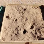 An outdoor wooden-framed sandbox filled with sand and partially covered by a green tarp, photographed from above