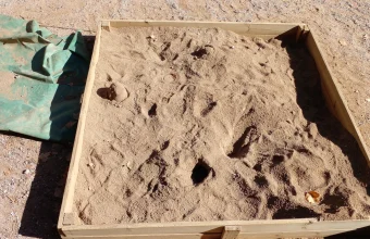 An outdoor wooden-framed sandbox filled with sand and partially covered by a green tarp, photographed from above