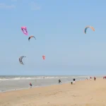Several colorful kites fly over a beach in De Haan, Belgium, as kitesurfers ride the waves below