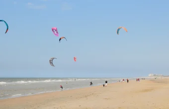 Several colorful kites fly over a beach in De Haan, Belgium, as kitesurfers ride the waves below