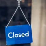 A blue Closed sign hanging inside a glass shop door
