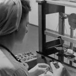 Black and white photo of a quality control inspector weighing a small precision part on an analytical balance scale