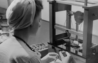 Black and white photo of a quality control inspector weighing a small precision part on an analytical balance scale