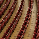 Rows of empty red theatre seats curving toward an empty stage, seen from above