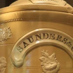 Close-up of an antique ceramic Saunders and Co. gravity water filter urn with ornate floral relief and a branded nameplate, Museum of Liverpool