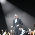 A conductor lit by a spotlight beam directs an orchestra on a dark stage, a visual metaphor for coordinating many automated systems from one governed point of control.