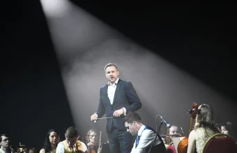 A conductor lit by a spotlight beam directs an orchestra on a dark stage, a visual metaphor for coordinating many automated systems from one governed point of control.