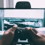 Close-up of hands holding a video game controller in front of a monitor displaying gameplay