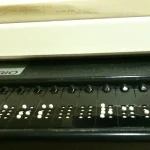 Close-up of a refreshable Braille display connected beneath a computer keyboard, showing raised dot patterns used by blind screen reader users