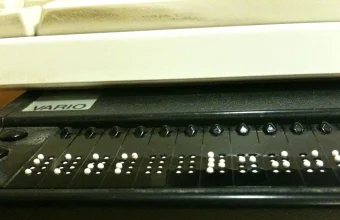 Close-up of a refreshable Braille display connected beneath a computer keyboard, showing raised dot patterns used by blind screen reader users